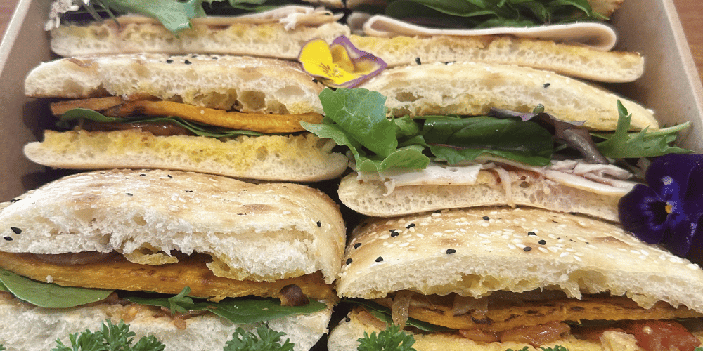 Close-up of multiple gourmet sandwiches filled with beef brisket, pickles and seeded mustard; roast chicken, pesto, red onion and lettuce; turkey, cranberry and brie; and semi-dried tomato, red onion, rocket and mozzarella, arranged in a cardboard tray. The rolls are topped with black sesame seeds and garnished with edible flowers.