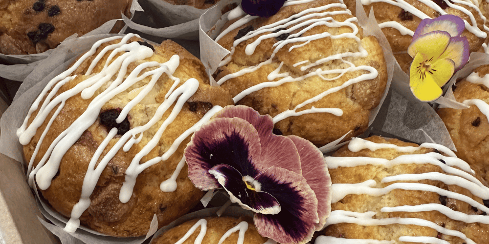 Close-up of several baked muffins in paper liners, drizzled with white icing and garnished with edible pansies. The pastries fill the frame, with one muffin visible in the top left corner.
