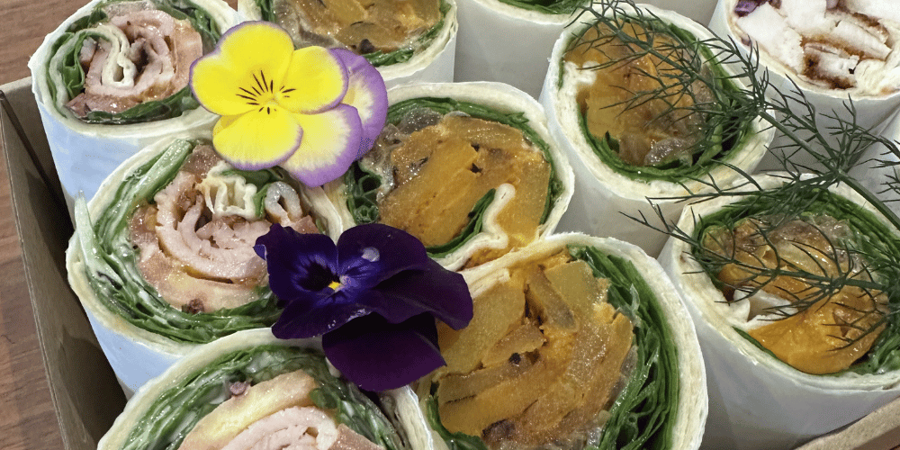 A close-up overhead view of several rolled wrap sandwiches arranged in a box, filled with chicken, lettuce, tomato and avocado; chicken schnitzel and slaw; smoked ham and salad; and turkey, cranberry and brie wraps
